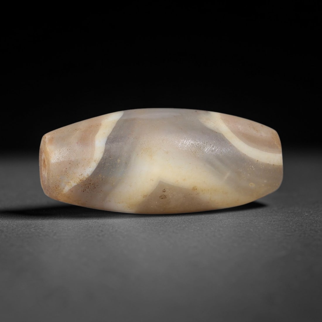 Ancient Banded Agate Bead - Silk Road Indus Valley Barrel Bead: Carved from banded agate, the bead is fashioned in an elongated barrel form, with a central drilled perforation for suspension. The stone exhibits striking natural banding in creamy white, soft taupe,