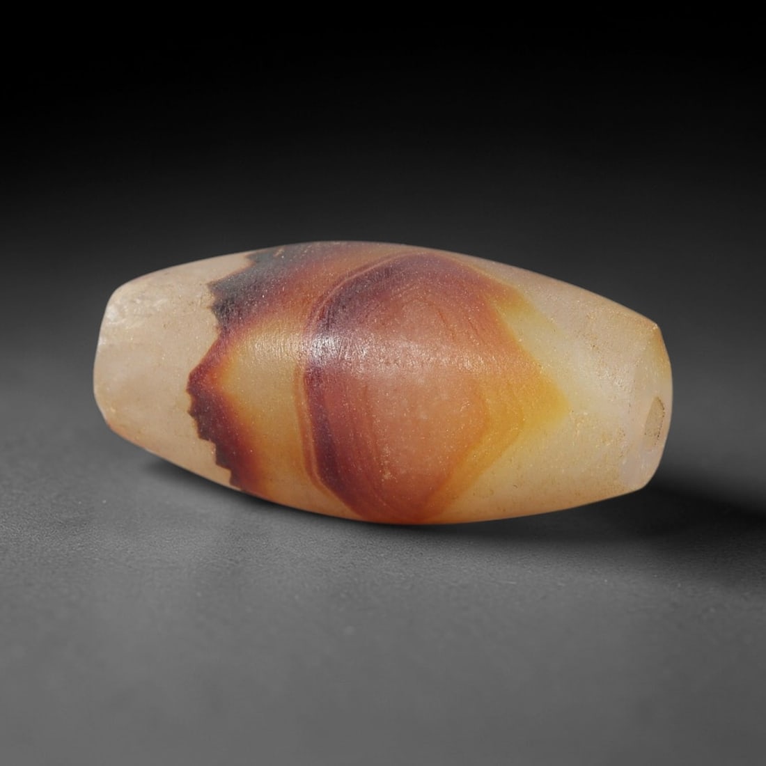 Ancient Agate Bead - Indus Valley Amulet: This artifact is crafted from variegated agate, exhibiting natural banding in warm amber, russet, and creamy white hues, shaped through careful lapidary work into a smooth barrel form. The bead featur