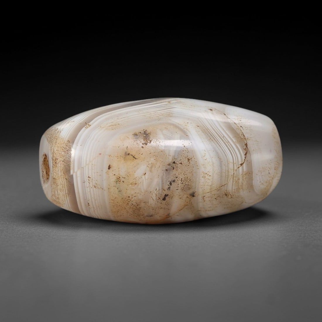 Ancient Banded Agate Bead - Central Asian Silk Road Barrel Bead: Carved from banded agate, the bead is fashioned in a compact barrel form, with a central drilled perforation for suspension. The stone exhibits delicate, wavy natural banding in milky white and soft b