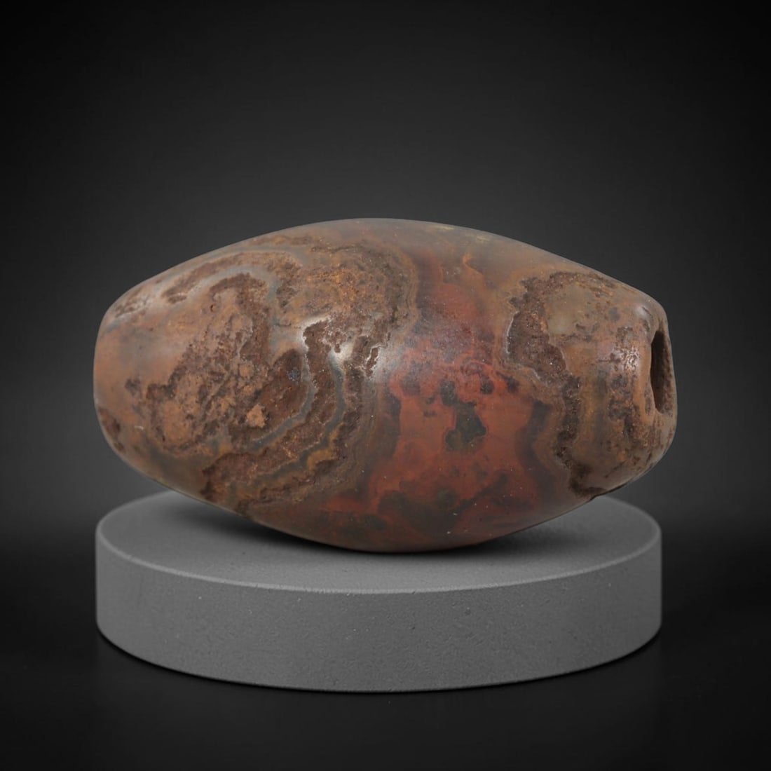 Ancient Jasper Bead - Silk Road Bactrian: Carved from banded jasper, the bead is fashioned in an ovoid barrel form with a central perforation for suspension. The stone displays rich natural concentric banding in deep reddish-brown, earthy och