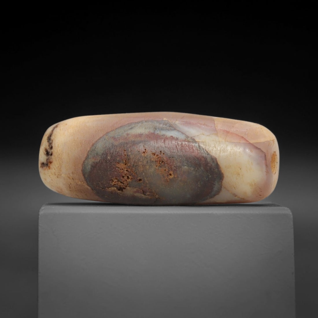 Ancient Agate Bead - Indus Valley Banded Carving: This artifact is crafted from banded agate, showcasing a striking, large oval inclusion of deep reddish-brown set against a soft, creamy pinkish-beige base across its polished surface. The bead is sha