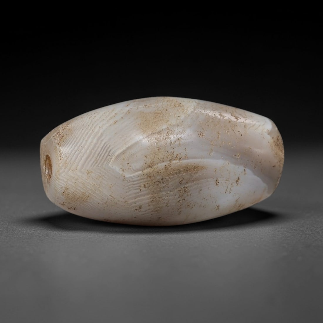 Ancient Agate Barrel Bead - Indus Valley Silk Road Bead: Carved from translucent banded agate, the bead is fashioned in a compact barrel form, with a central drilled perforation for suspension. The stone exhibits fine, wavy natural banding in milky white an