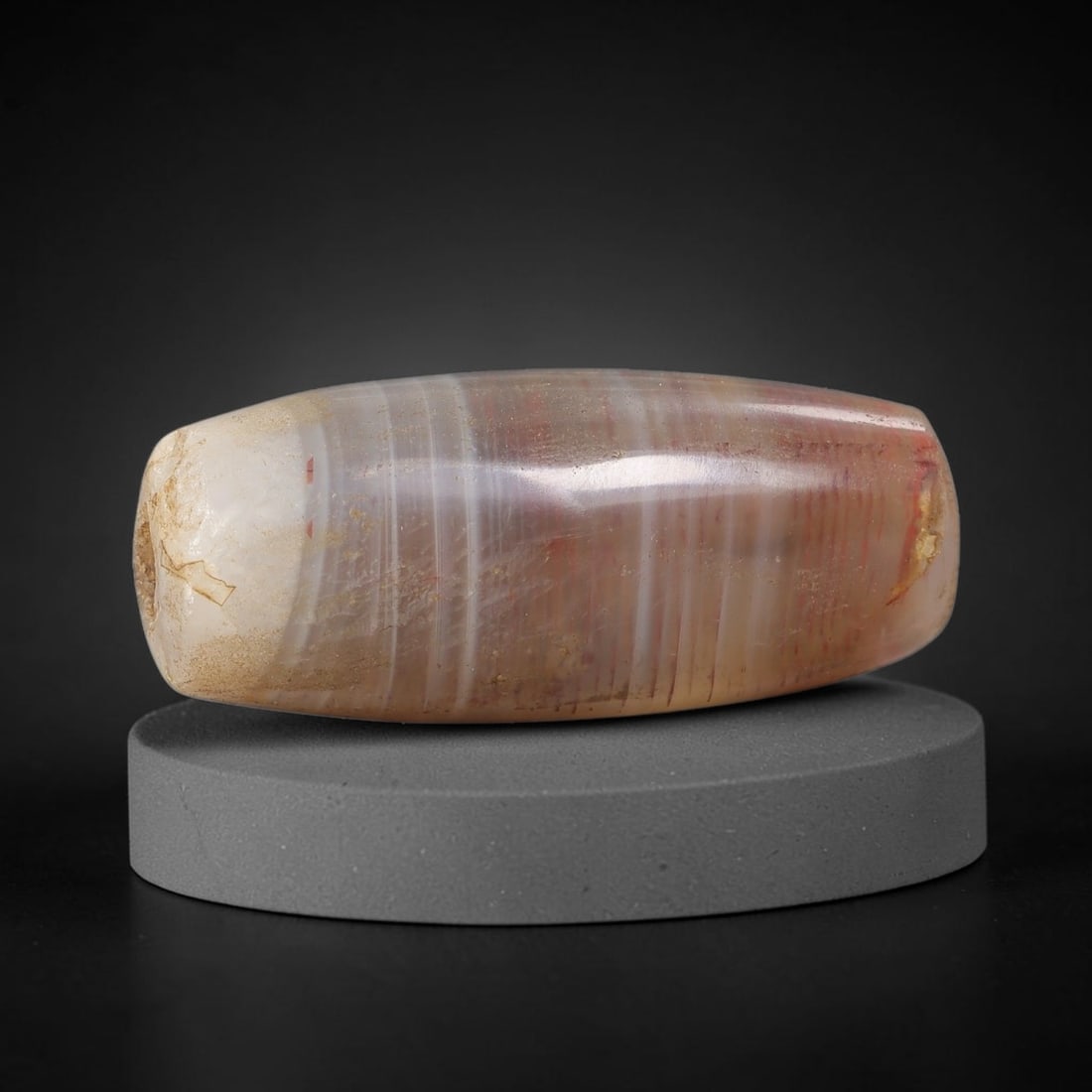 Ancient Banded Agate Bead - Indus Valley Artifact, 59BC: This artifact is crafted from natural banded agate, shaped into an elongated barrel form with fine parallel concentric banding in soft taupe, white, and subtle red hues. The bead features a central pe