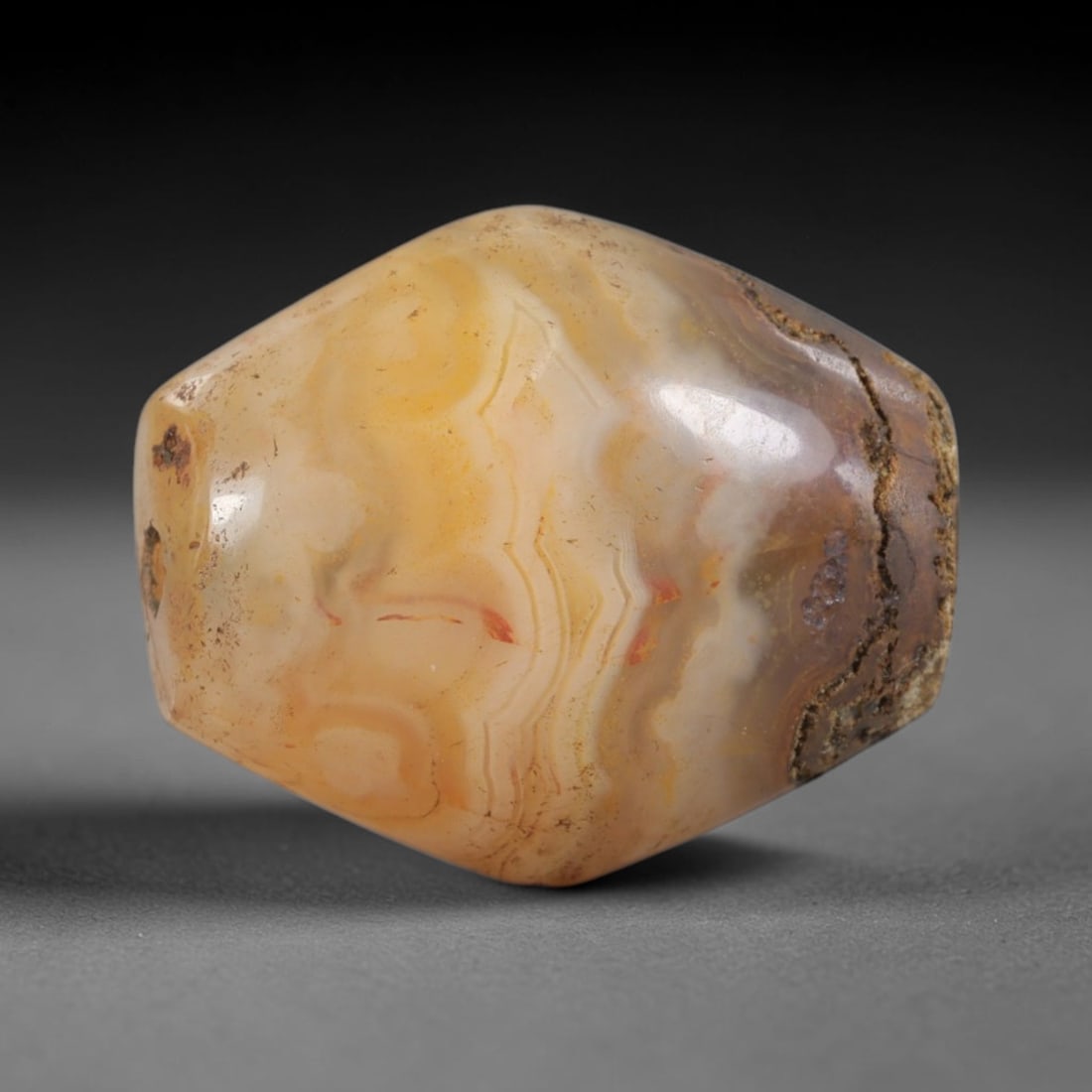 Ancient Agate Bead - Indus Valley Banded Carving: This artifact is crafted from banded agate, showcasing natural concentric banding in warm amber, cream, and earthy brown tones. The bead is shaped in a faceted, barrel form with a central perforation