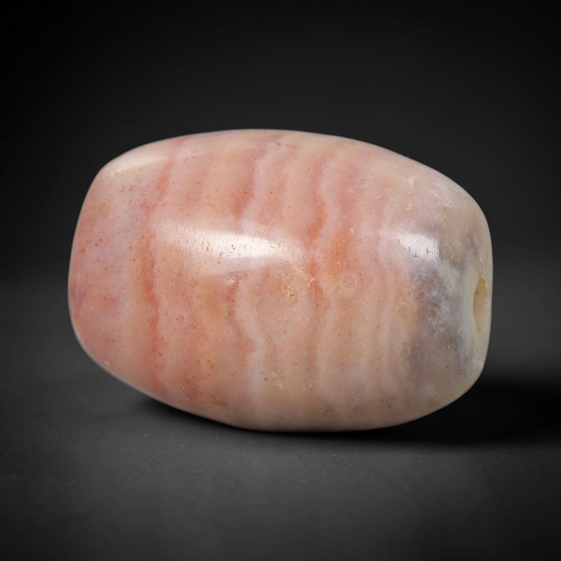 Ancient Pink Agate Bead - Silk Road Indus Valley Barrel Bead: Carved from banded agate, the bead is fashioned in a compact barrel form, with a central drilled perforation for suspension. The stone exhibits delicate, wavy natural banding in soft blush pink and pa