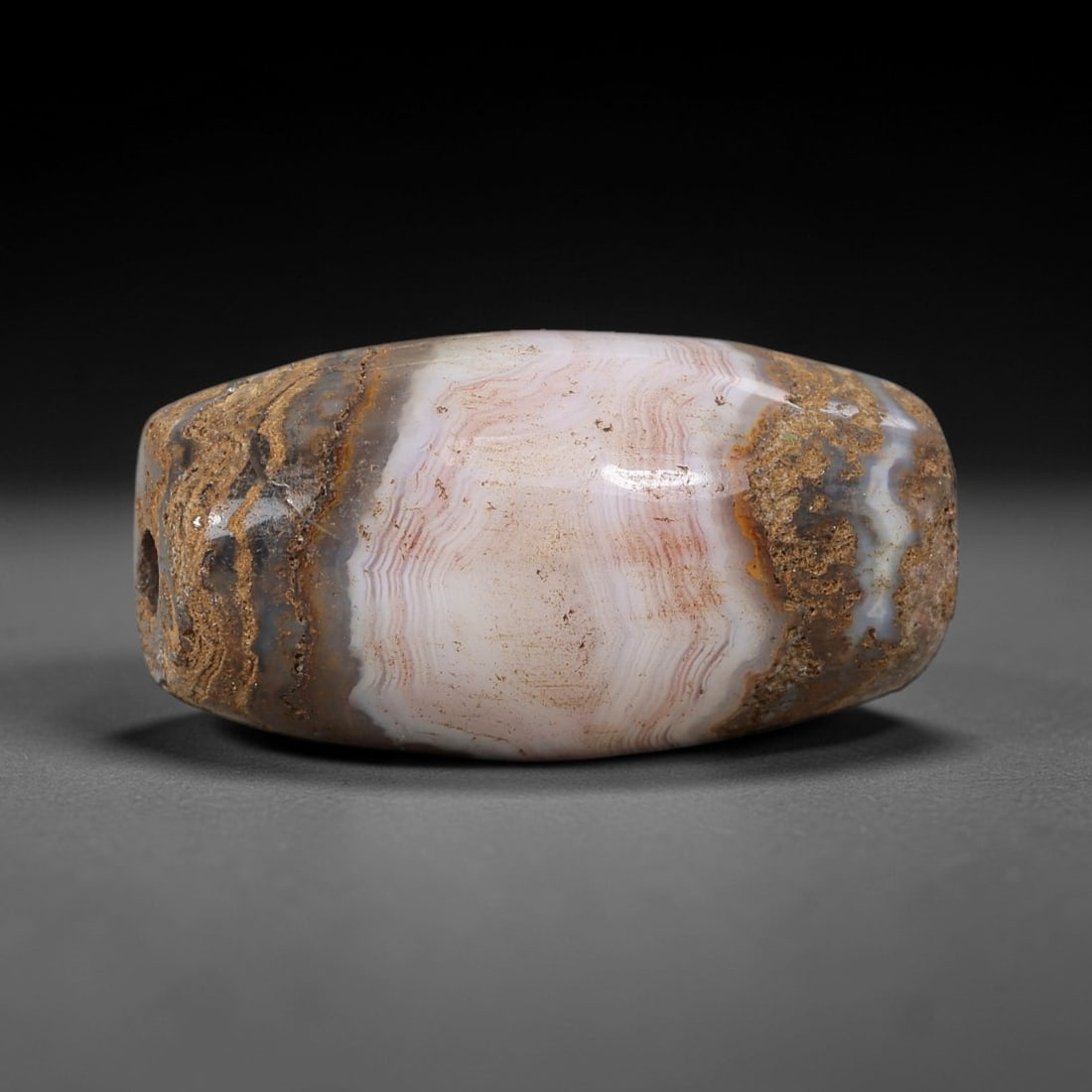 Ancient Banded Agate Bead - Silk Road Indus Valley Barrel Bead: Carved from banded agate, the bead is fashioned in a compact barrel form, with a central drilled perforation for suspension. The stone exhibits striking natural banding in soft pink, creamy white, and