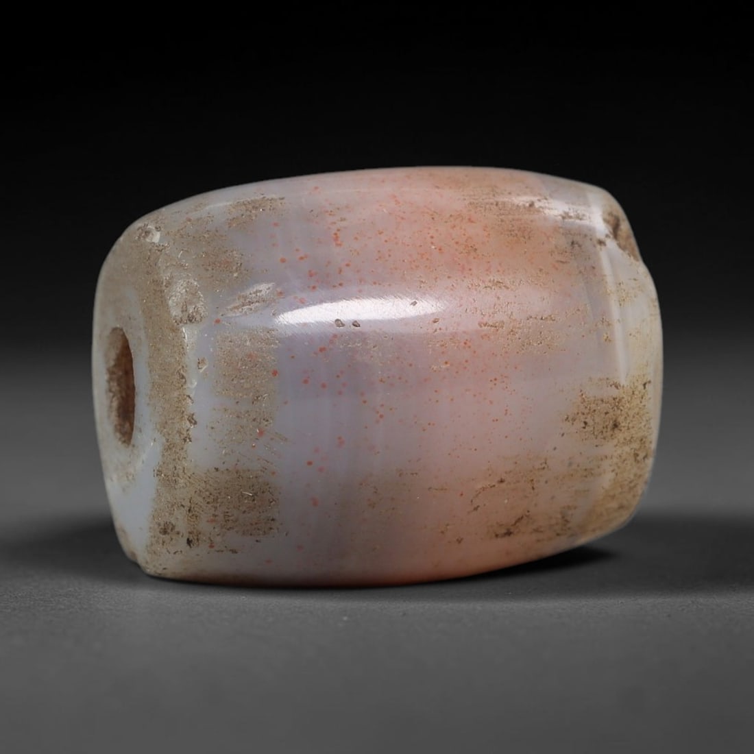 Ancient Agate Bead - Indus Valley Amulet: This artifact is crafted from pale pinkish agate, exhibiting subtle banding and scattered vivid red mineral inclusions across its smooth barrel form, shaped through skilled ancient lapidary work. The