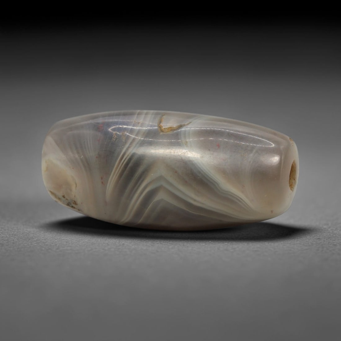 Ancient Agate Bead - Indus Valley Banded Carving: This artifact is crafted from banded agate, exhibiting striking chevron-style concentric banding in translucent grey, creamy white, and pale amber tones across its polished, faceted surface. The bead