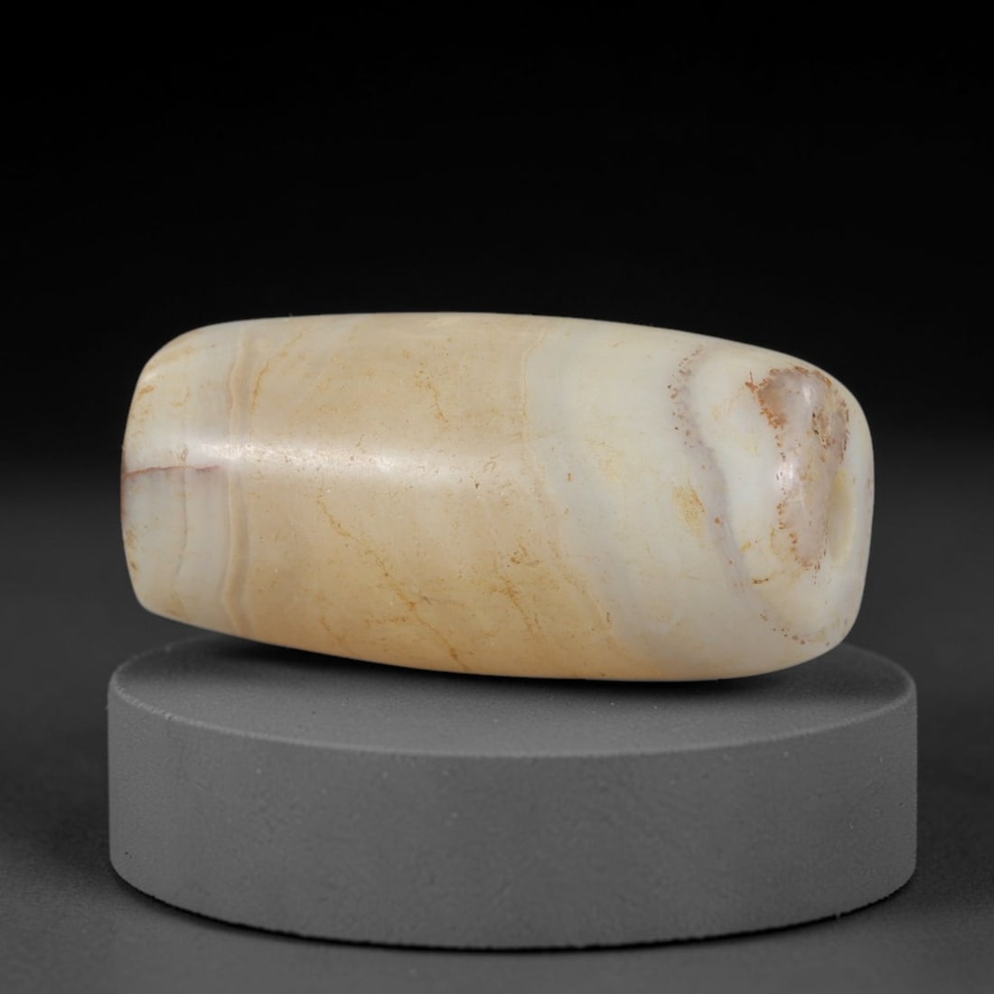 Ancient Agate Bead - Indus Valley Silk Road: Carved from banded agate, the bead is fashioned in a cylindrical barrel form with a central perforation for suspension. The stone displays natural concentric banding in creamy beige, pale grey, and so