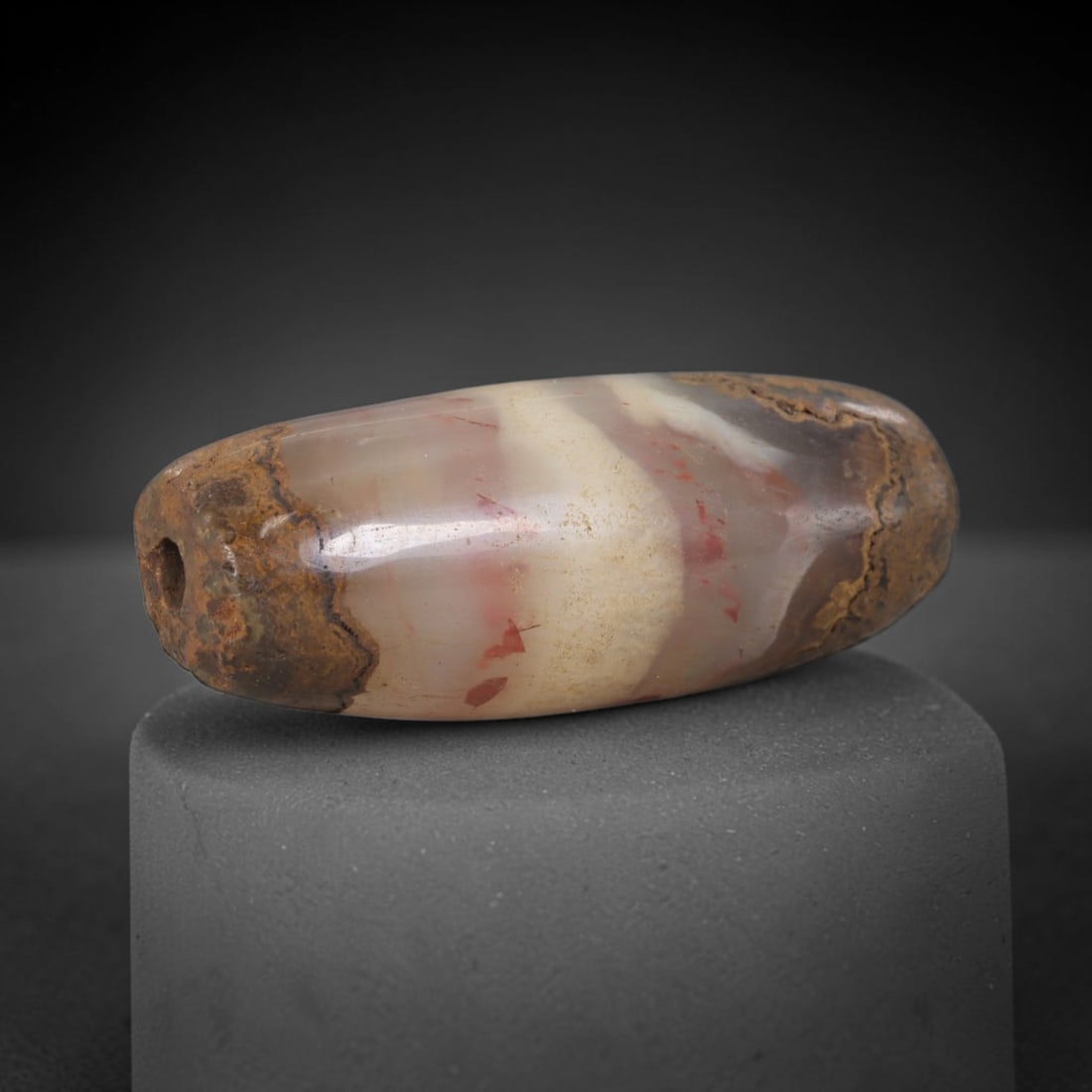 Ancient Agate Bead - Silk Road Bactrian: Carved from banded agate with natural red inclusions, the bead is fashioned in a cylindrical barrel form with a central perforation for suspension. The stone displays flowing concentric banding in cre