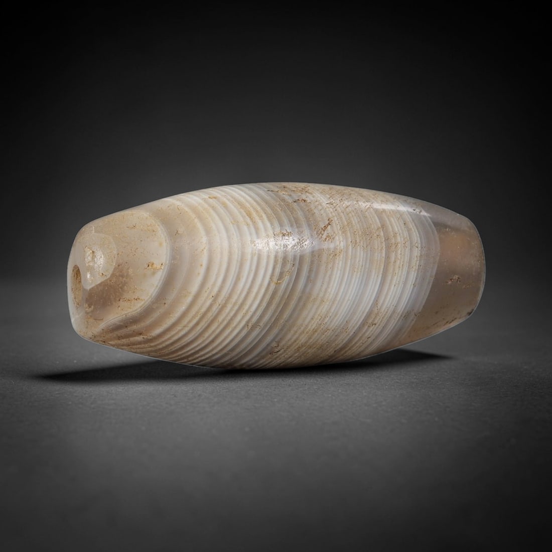 Ancient Banded Agate Bead - Silk Road Indus Valley Barrel Bead: Carved from translucent banded agate, the bead is fashioned in a classic barrel form, with a central drilled perforation for suspension. The stone exhibits striking, fine concentric banding in creamy