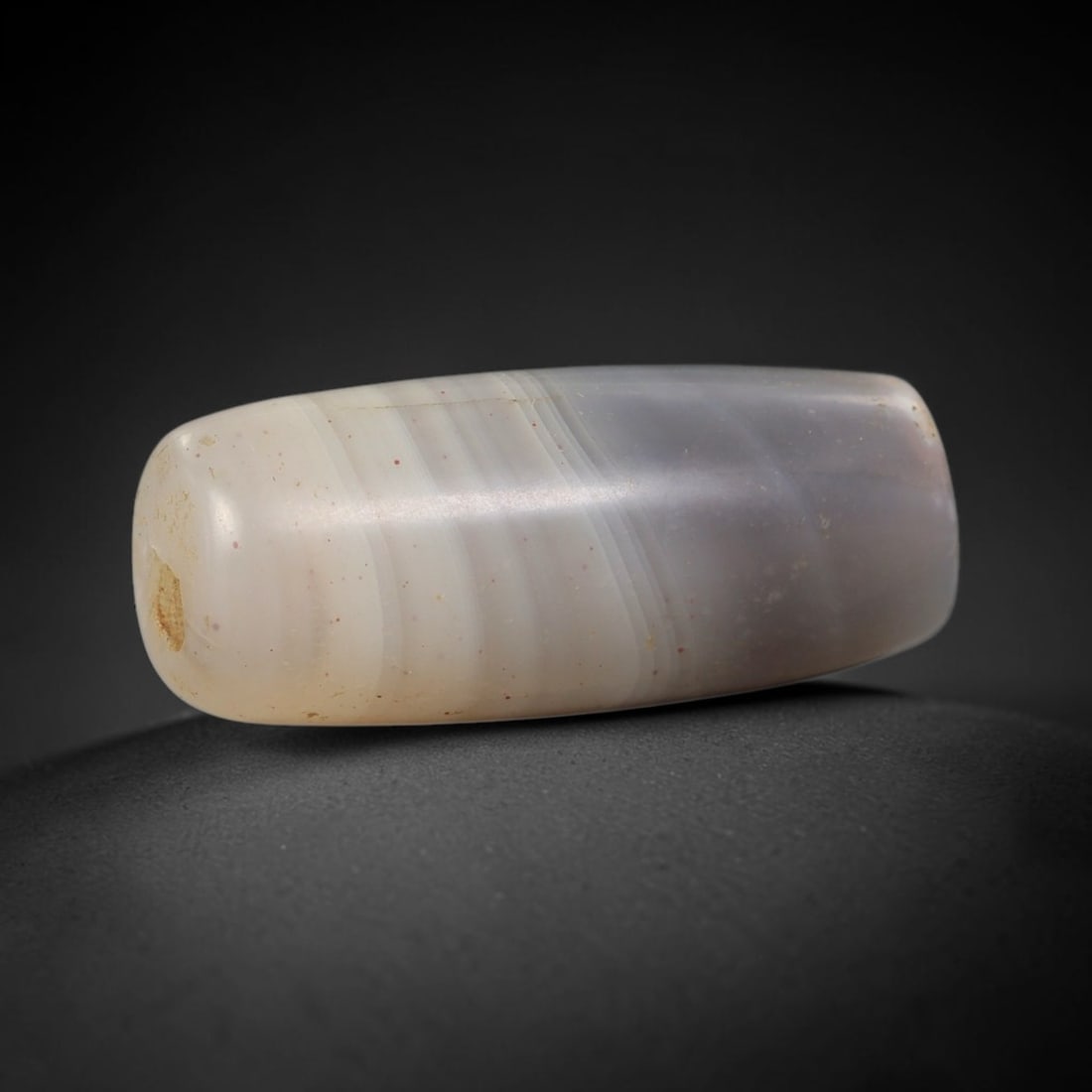 Ancient Banded Agate Bead - Indus Valley Artifact: This artifact is crafted from natural banded agate, shaped into an elongated barrel form with delicate, parallel concentric banding in soft grey and cream hues, accented by tiny red mineral specks. Th