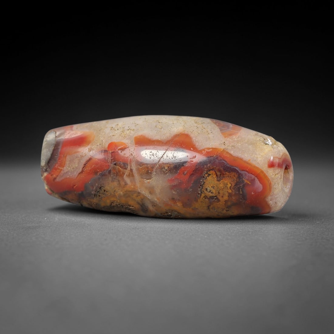 Ancient Agate Bead - Tibetan Amulet: This artifact is crafted from richly variegated agate, displaying bold bands of vivid carmine red, amber, creamy white, and earthy brown mineral inclusions, shaped into a smooth barrel form through sk