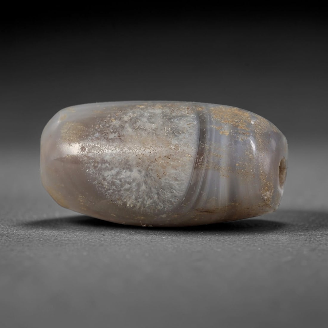 Ancient Agate Bead - Indus Valley Artifact: This artifact is crafted from banded agate, shaped into a smooth, elongated barrel form with natural grey and cream banding and distinctive dendritic mineral inclusions. The bead features a central pe