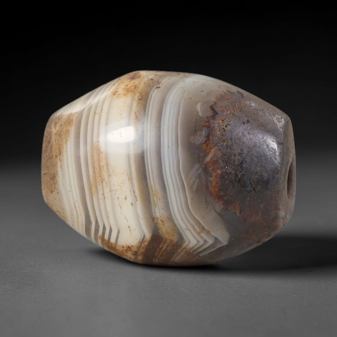 Ancient Agate Bead - Indus Valley Carving: This artifact is crafted from banded agate, exhibiting fine, concentric banding patterns characteristic of ancient stoneworking techniques. The form is a barrel-shaped bead with a smooth, polished sur