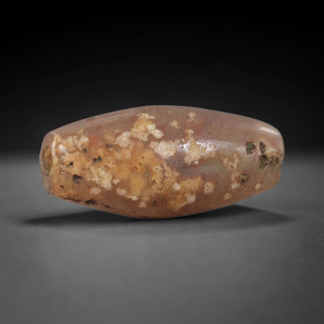 Ancient Agate Bead - Silk Road Western Asiatic: This artifact is fashioned from natural agate, displaying a translucent base with inclusions of white mineral deposits and earthy patination across its surface. The bead is shaped in an elongated, bic