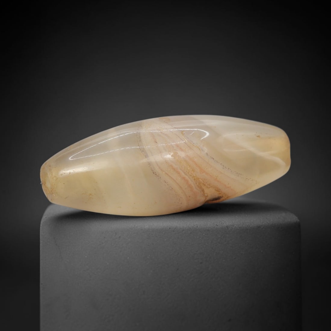 Ancient Agate Bead - Indus Valley Banded Carving: This artifact is crafted from banded agate, exhibiting soft, flowing concentric banding in warm honey-amber and creamy translucent tones across its polished, elongated bicone surface. The bead is shap