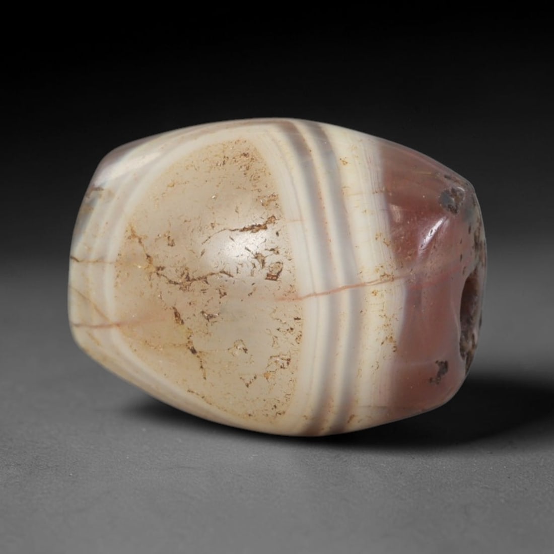 Ancient Agate Bead - Silk Road Bactrian: Crafted from banded agate, the bead is formed in an ovoid barrel shape with a central perforation for suspension. The stone features distinct concentric banding in creamy off-white, pale greyish-brown