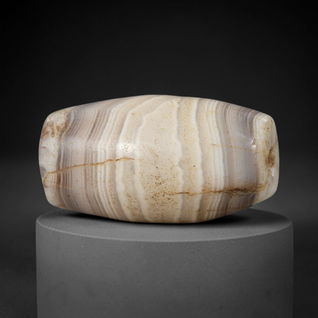 Ancient Agate Bead - Near Eastern Amulet: This artifact is crafted from banded agate, showcasing fine concentric layered striations in creamy white, soft grey, and warm beige tones, shaped into a smooth barrel form through skilled ancient lap