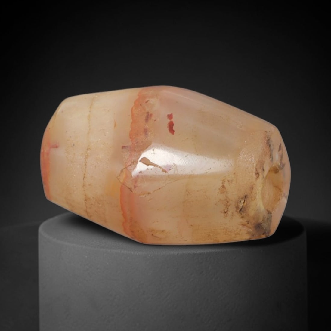 Ancient Agate Bead - Silk Road Western Asiatic: This artifact is fashioned from translucent agate, featuring subtle natural banding and faint red mineral inclusions across its surface. The bead is shaped in a faceted, barrel form with a central per