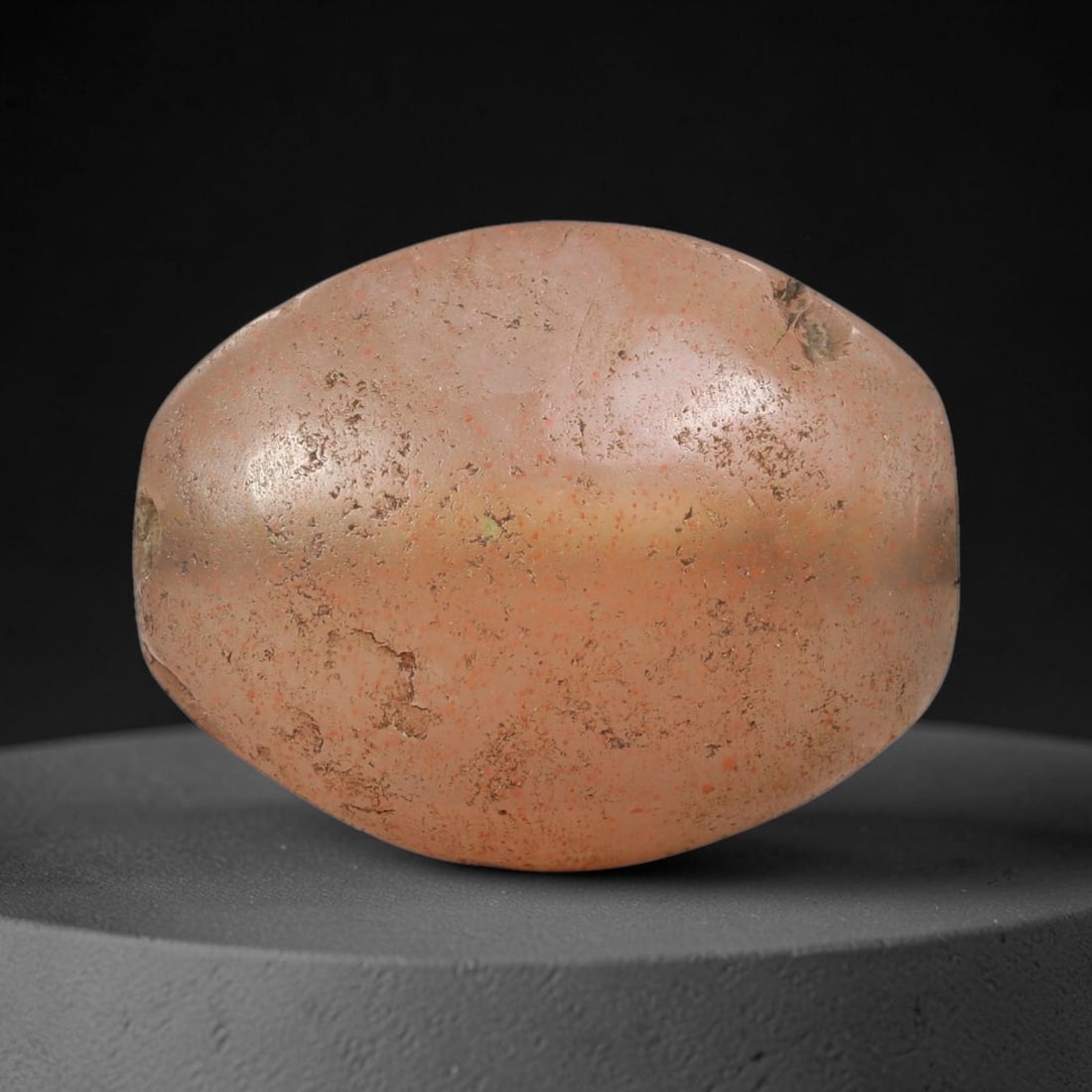 Ancient Agate Bead - Indus Valley Artifact: This artifact is crafted from translucent agate, shaped into a large, rounded oval (barrel) form with a warm peachy-pink base and scattered red mineral inclusions. The bead features a central perforat