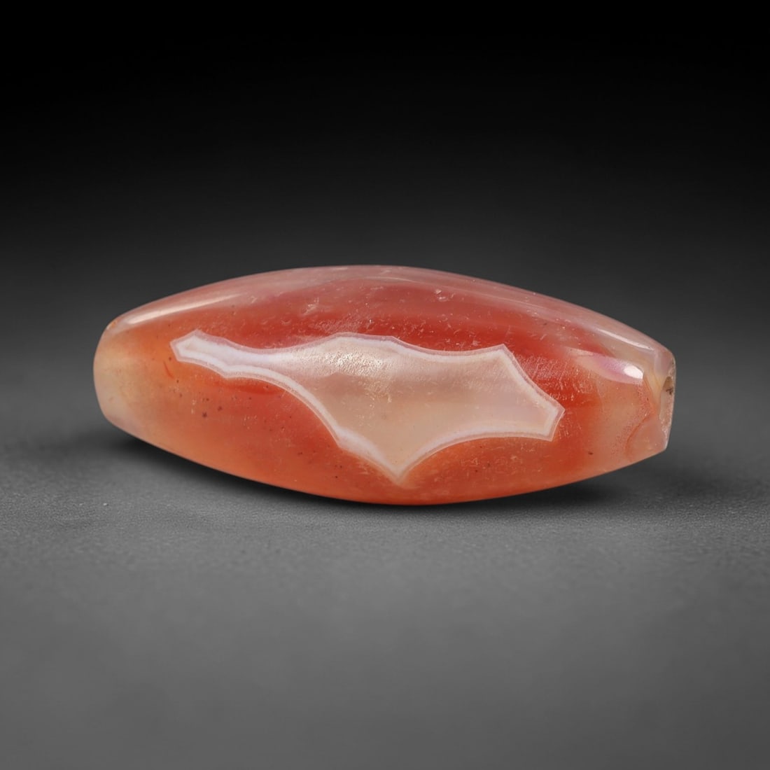 Ancient Carnelian Agate Bead - Silk Road Indus Valley Bicone Bead: Carved from translucent banded carnelian agate, the bead is fashioned in a slender bicone form, with a central drilled perforation for suspension. The stone displays a striking natural banding pattern