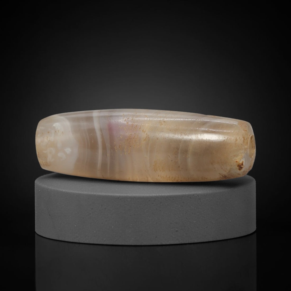 Ancient Agate Bead - Indus Valley Banded Carving: This artifact is crafted from banded agate, exhibiting subtle, flowing concentric banding in warm cream, pale amber, and translucent grey tones across its polished, elongated surface. The bead is shap