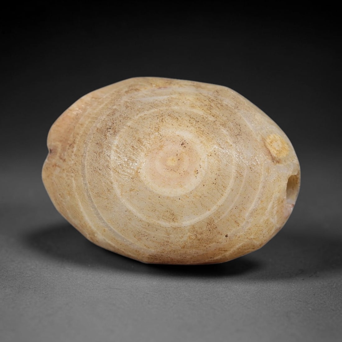 Ancient Agate Bead - Indus Valley Artifact: This artifact is carved from banded agate, shaped into a flattened ovoid form with a striking concentric circular banding pattern radiating from a central core. The bead features a lateral perforation