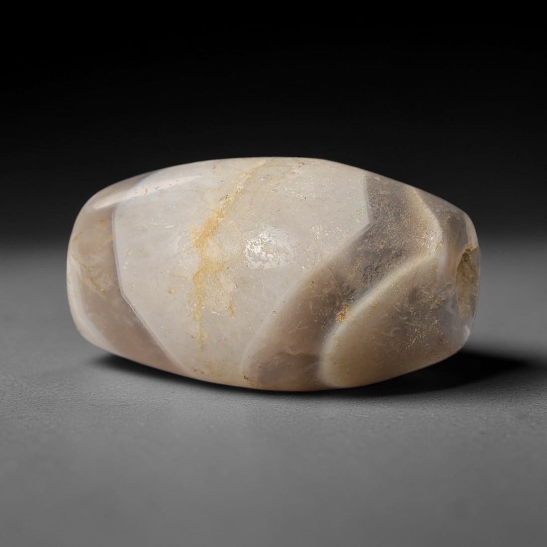 Ancient Agate Bead - Bactrian Amulet: This artifact is crafted from banded agate, exhibiting layered tones of pale grey, creamy white, and soft brown, shaped into a faceted barrel form through skilled ancient lapidary work. The bead featu