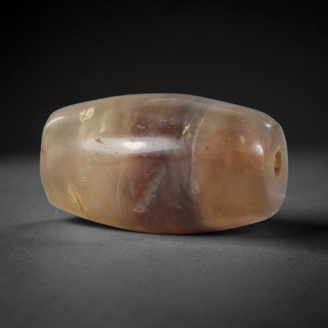 Ancient Agate Bead - Silk Road Bactrian: Carved from banded agate, the bead is fashioned in a cylindrical barrel form with a central perforation for suspension. The stone displays natural, muted concentric banding in creamy beige, translucen