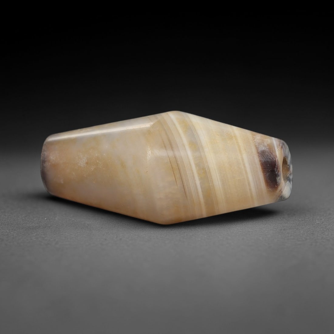 Ancient Agate Bead - Silk Road Indus Valley Bead: Carved from banded agate, the bead is fashioned in a distinctivebarrel form, with a central drilled perforation for suspension. The stone displays natural concentric banding in creamy beige, pale grey