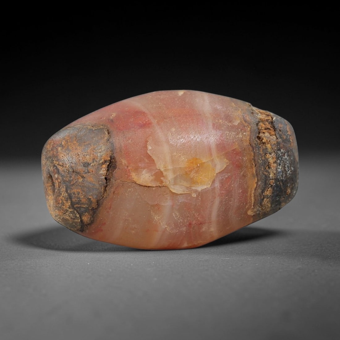 Ancient Carnelian Agate Bead - Silk Road Indus Valley Barrel Bead: Carved from banded carnelian agate, the bead is fashioned in a compact barrel form, with a central drilled perforation for suspension. The stone exhibits natural banding in soft pink, warm orange, and