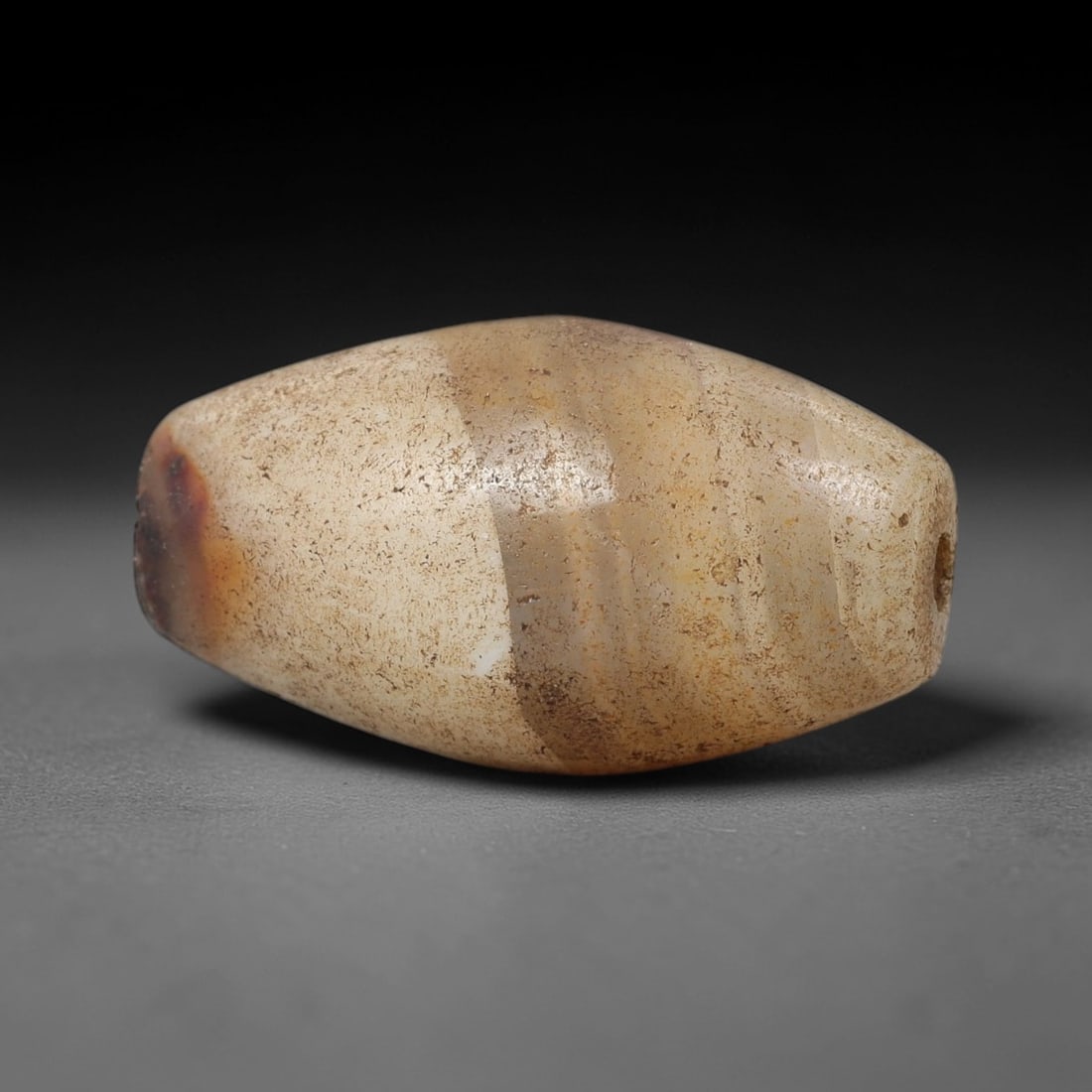 Ancient Agate Barrel Bead - Indus Valley Silk Road Bead: Carved from banded agate, the bead is fashioned in a compact barrel form, with a central drilled perforation for suspension. The stone exhibits natural banding in creamy white, warm tan, and deep ambe
