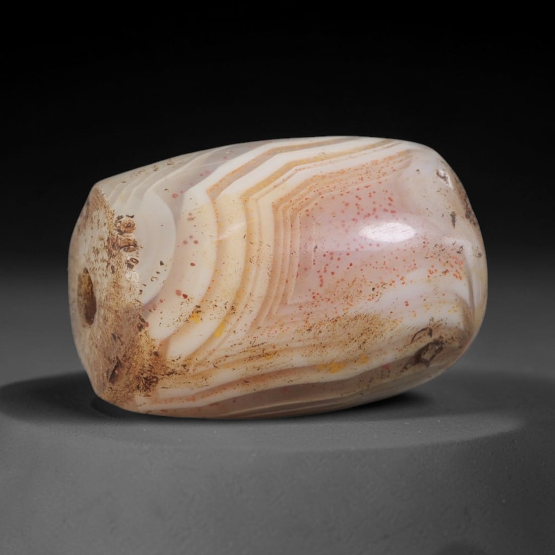 Ancient Agate Bead - Indus Valley Banded: This artifact is crafted from banded agate, exhibiting prominent, layered concentric banding in creamy white and warm amber hues, accented by fine red mineral speckling across the polished surface. Th