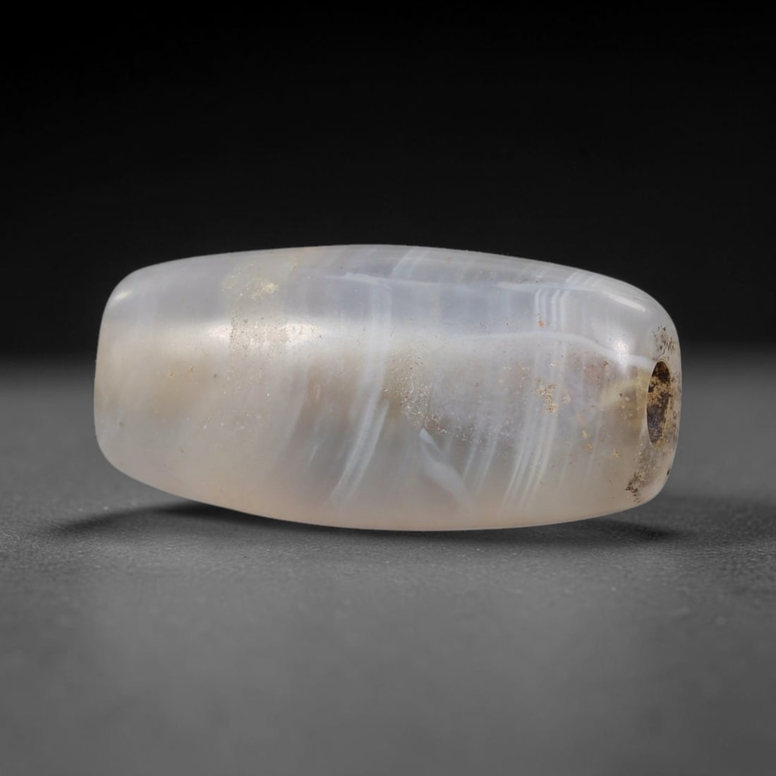 Ancient Agate Bead - Near Eastern Artifact: This artifact is crafted from translucent banded agate, shaped into a smooth, elongated barrel form with delicate wavy white banding across its surface. The bead features a central perforation for sus
