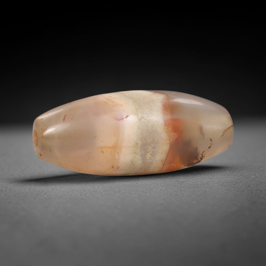 Ancient Agate Bead - Afghan Amulet: This artifact is crafted from translucent agate, displaying delicate banding in soft peach, pale amber, and creamy white tones with subtle mineral veining, shaped into a smooth elongated barrel form t
