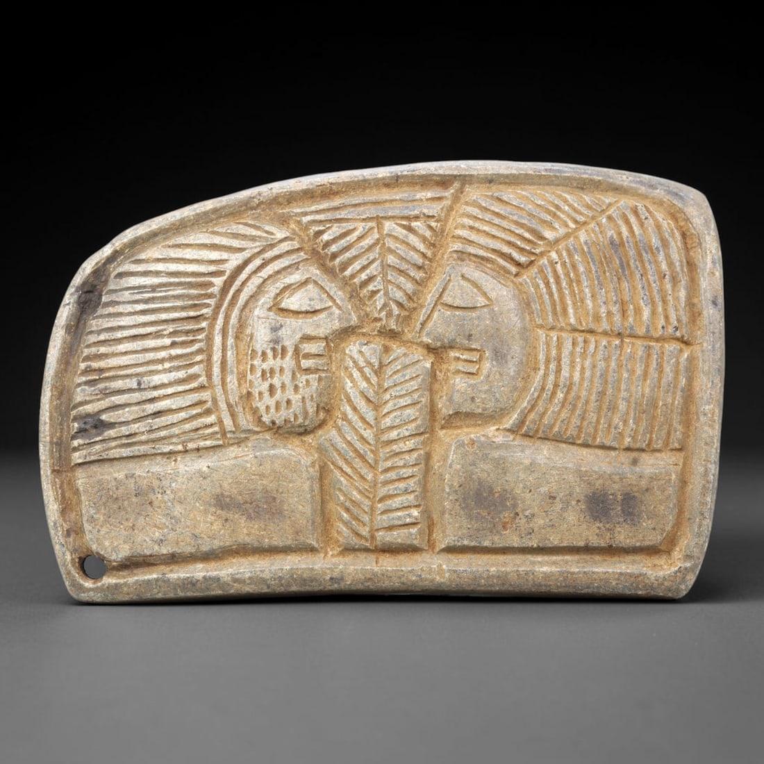 Ancient Stone Plaque Amulet - Near Eastern Dual Human Figural Plaque, 12ZY: Carved from a single piece of stone, this rectangular relief amulet depicts two stylized human figures in profile facing one another, flanking a central stylized tree motif, rendered in the linear, in