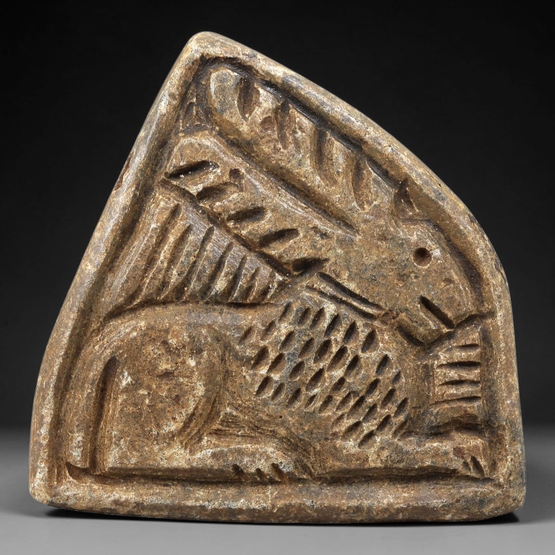 Ancient Stone Plaque Relief - Near Eastern Winged Ibex Figurine, 67BE: Carved from a single piece of stone, this triangular relief plaque depicts a recumbent winged ibex, rendered in the stylized, linear artistic tradition of the ancient Near East. The figure is detailed