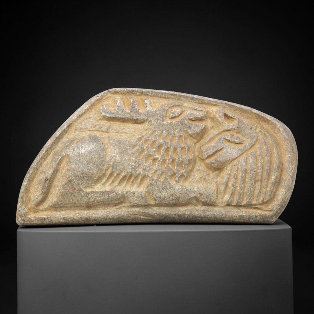 Ancient Stone Plaque Relief - Near Eastern Sphinx & Feline Figural Plaque, 13ZY: Carved from a single piece of stone, this curved relief plaque depicts a stylized sphinx (a composite mythological creature with a lion's body and human head) facing a feline, rendered in the linear,