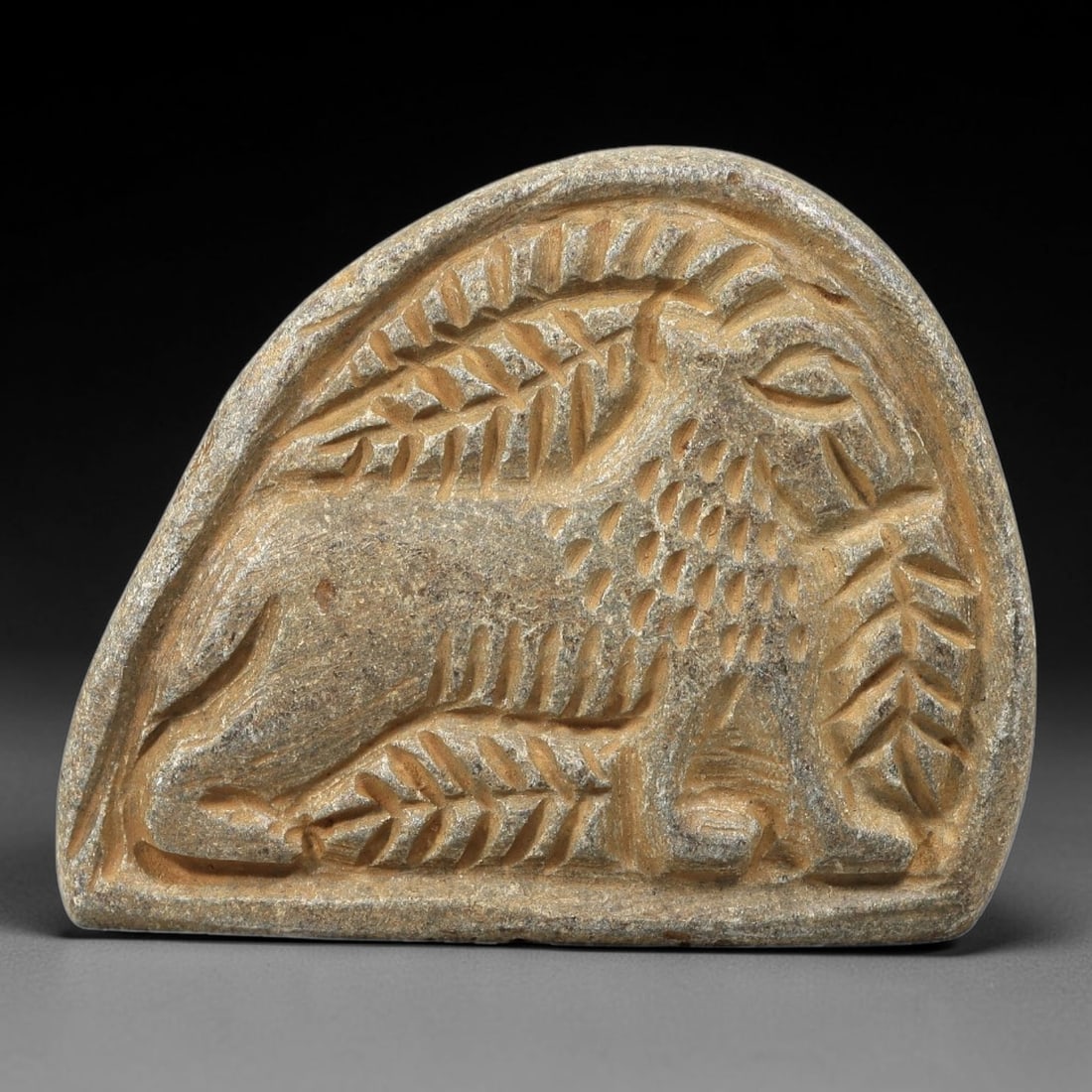 Ancient Stone Plaque Relief - Near Eastern Ibex Figurine, 64BE: Carved from a single piece of stone, this semi-circular relief plaque depicts a recumbent ibex, rendered in the stylized, linear artistic tradition of the ancient Near East. The figure is detailed wit
