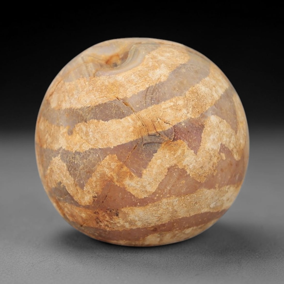 Ancient Carnelian Etched Bead - Gandhara Archaeological Find, 25BD: Carved from natural carnelian, the bead is spherical in form, adorned with white etched zigzag and linear band motifs across its surface. A lateral drilled suspension hole is present, characteristic o