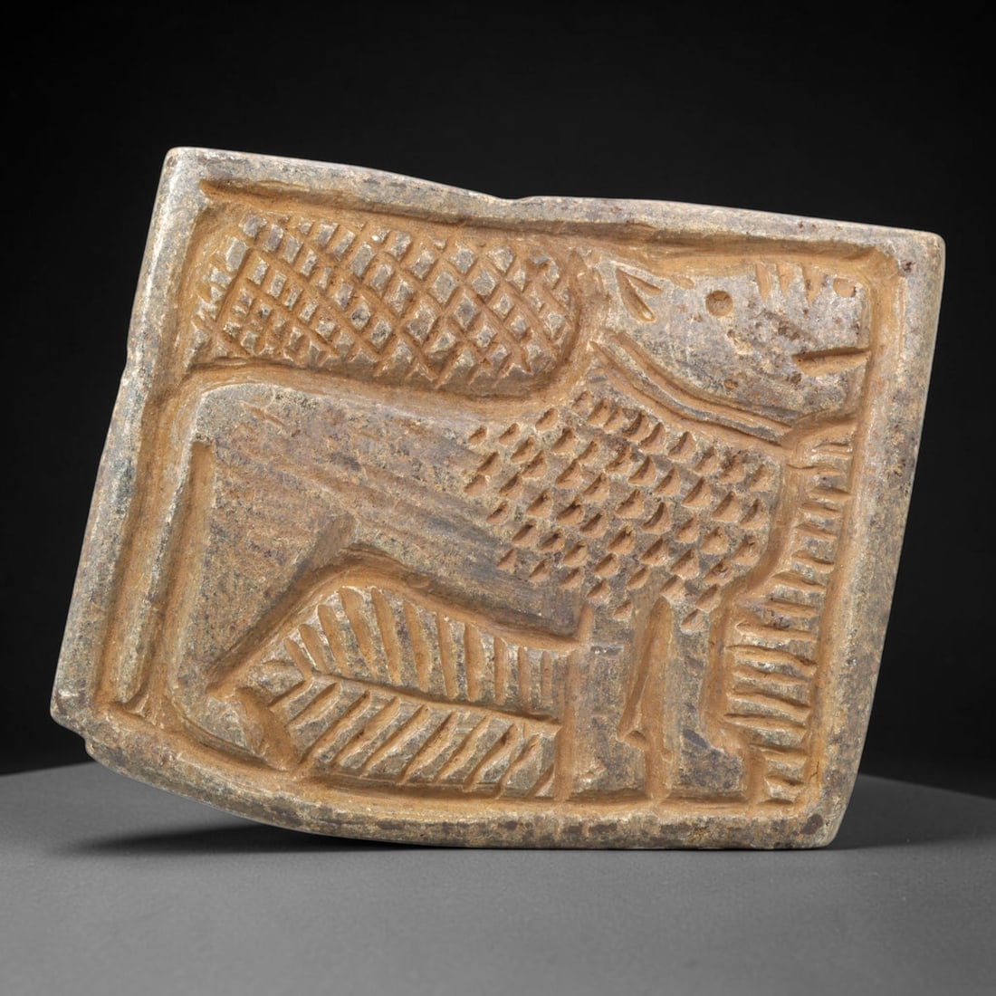 Ancient Stone Plaque Relief - Near Eastern Feline Figurine, 17ZY: Carved from a single piece of stone, this rectangular relief plaque depicts a recumbent feline, rendered in the stylized, linear artistic tradition of the ancient Near East. The figure is detailed wit