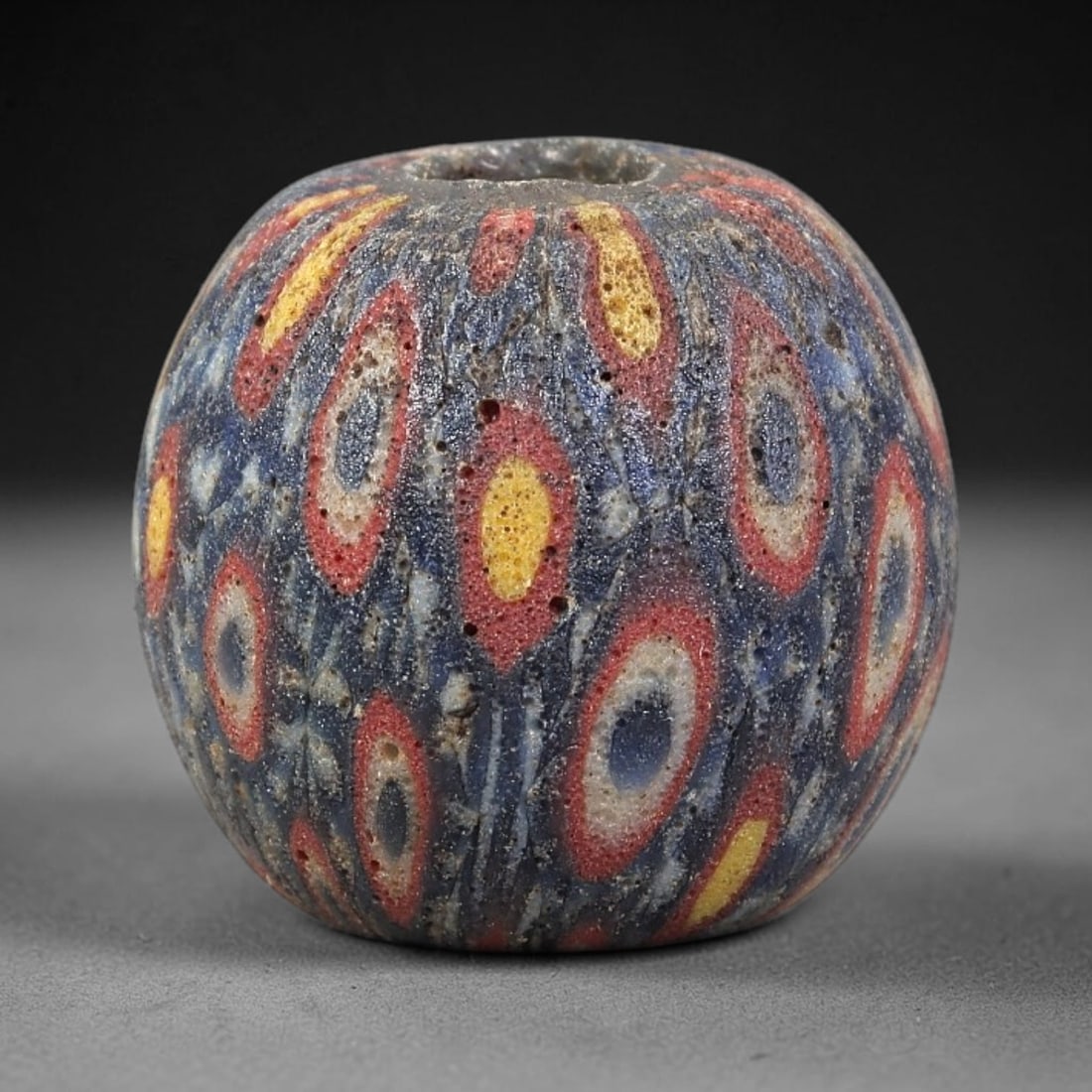 Ancient Glass Bead - Roman Millefiori Spherical Pendant, 44BG: A spherical ancient glass bead crafted in the millefiori mosaic technique, with a deep blue glass base overlaid with intricate fused cane patterns. The surface is decorated with repeating eye-like mot