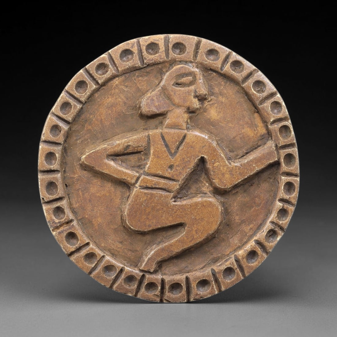 Ancient Steatite Plaque - Near Eastern Human Figure Relief, 89BE: Carved from steatite, the circular plaque features a raised relief of a human figure in a dynamic kneeling or dancing pose, rendered with a stylized head, V-neck tunic, and defined waistband. The figu