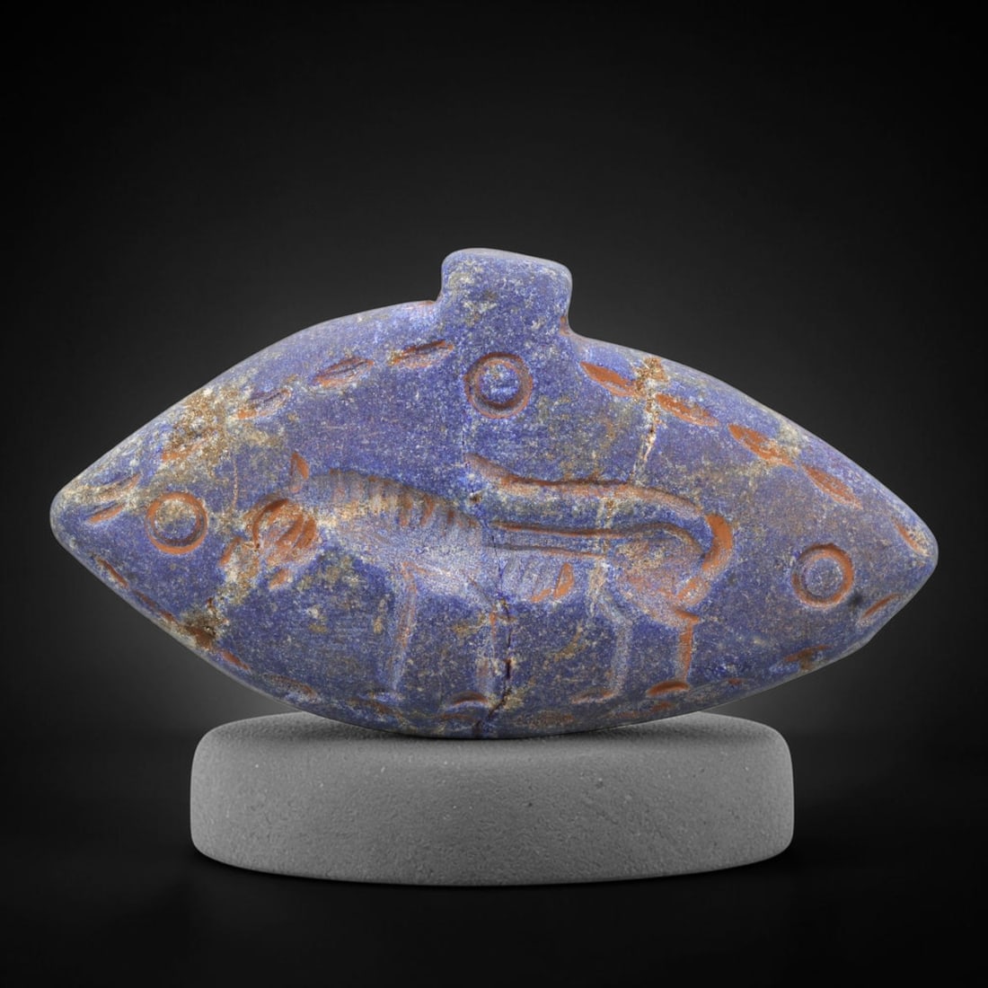 Ancient Lapis Amulet - Near Eastern Animal Figure, 76ZY: Carved from lapis lazuli, the amulet is shaped as a pointed oval, incised with a stylized quadrupedal animal figure surrounded by circular and linear decorative motifs, with reddish pigment filling th