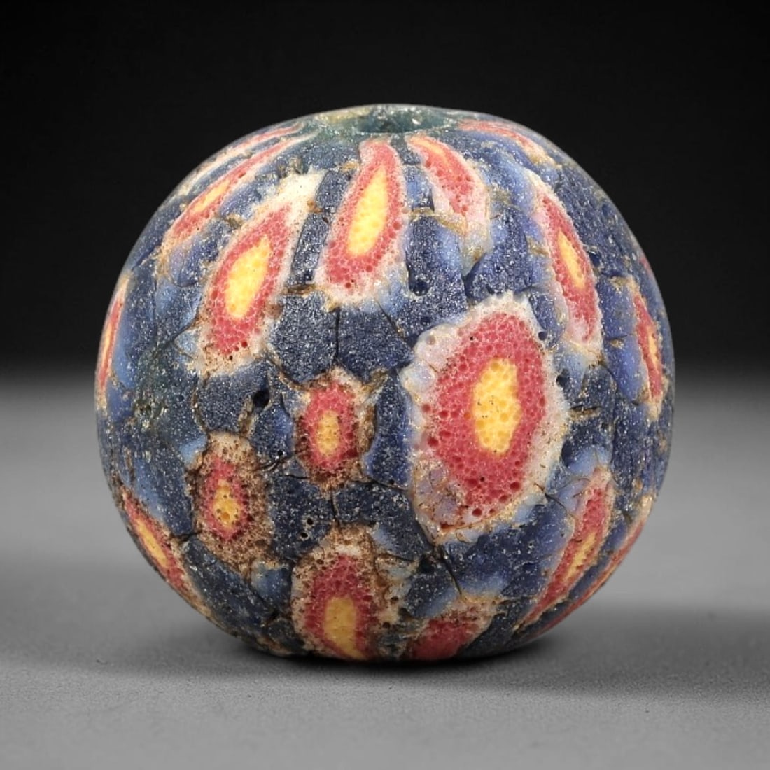 Ancient Glass Bead - Roman Millefiori Spherical Pendant, 23BG: A spherical ancient glass bead crafted in the millefiori mosaic technique, with a deep blue glass base overlaid with intricate fused cane patterns. The surface is decorated with repeating teardrop-sha