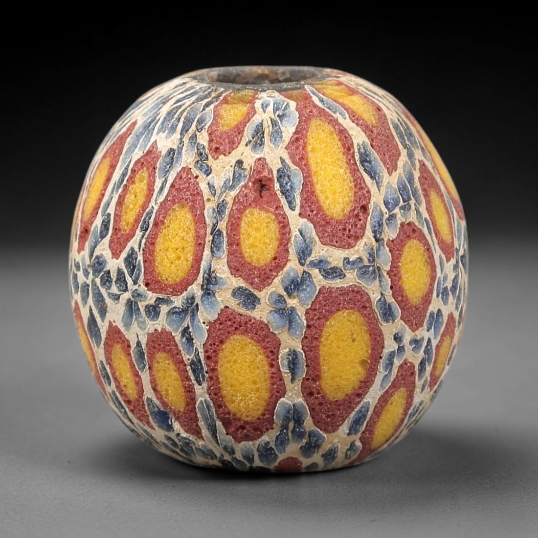 Ancient Glass Bead - Roman Millefiori Spherical Pendant, 27BG: A spherical ancient glass bead crafted in the millefiori mosaic technique, with a creamy white glass base overlaid with intricate fused cane patterns. The surface is decorated with repeating oval moti