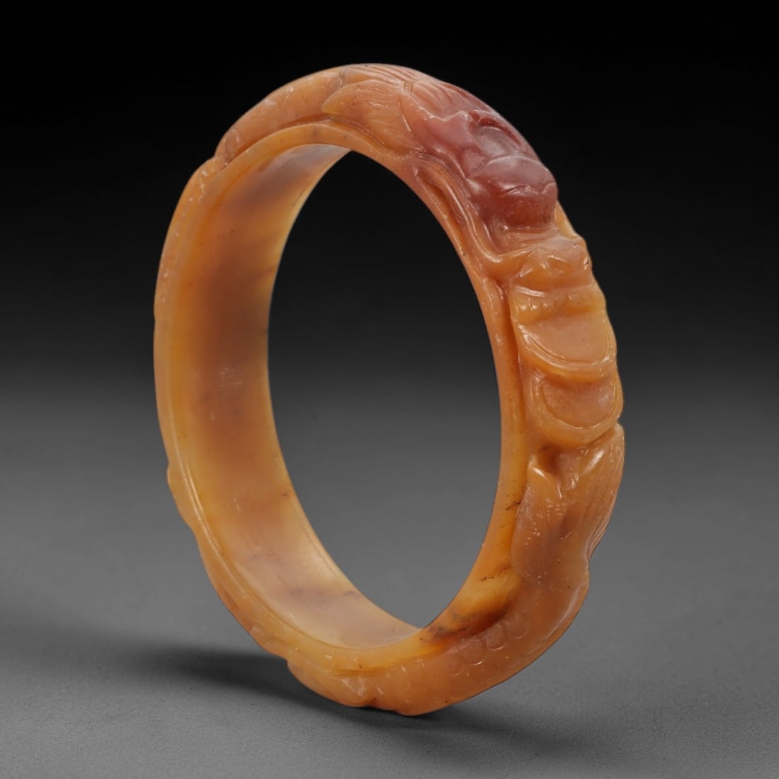 A Burmese Jadeite Jade Bangle, 20th Century, 67ZL: Carved from pale yellowish-orange jadeite jade with natural reddish inclusions distributed throughout the matrix, the bangle is formed as a circular band with a raised, relief-carved dragon motif on t
