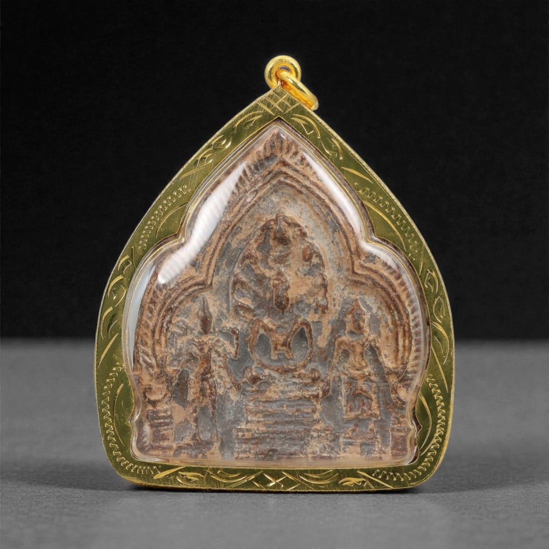A Thai Buddhist Composite Amulet in Gilt Metal Frame, 20th Century, 51FN: The amulet is encased in a heart-shaped gilt metal frame with fine incised linear border detailing and a suspension loop at the apex. The obverse features a composite plaque of compressed organic mate