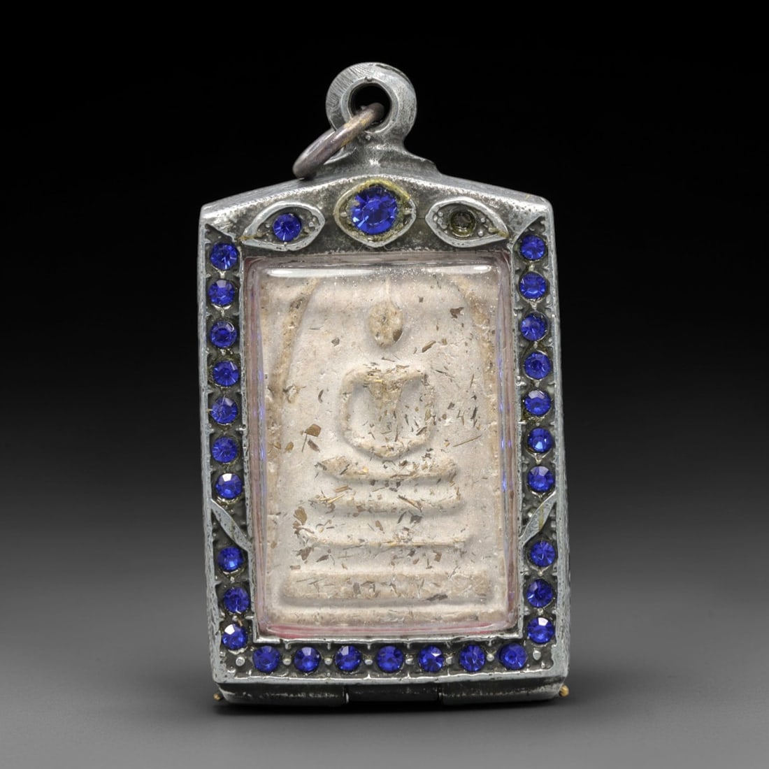 A Thai Phra Somdet Clay Amulet in a Gem-Set Silver-Tone Case, 20th Century, 13ZK: This rectangular Phra Somdet amulet is crafted from sacred beige clay, embedded with fine organic inclusions, and encased in an ornate silver-tone metal case with a suspension loop at the apex. The ob